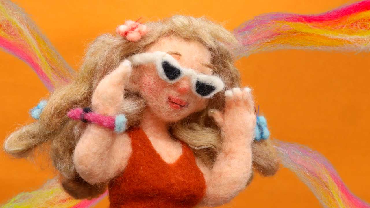 A felted wool doll with long blond hair, wearing white sunglasses, a red top, and colorful bracelets poses against an orange background with rainbow streaks, inspired by the Coachella 2026 visual identity film by Andrea Love and Hornet. Frame from Stash Magazine article.