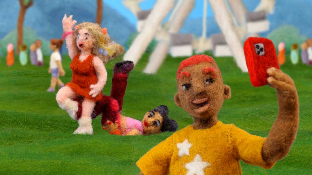 Three felted wool figures in a park: a person in a yellow shirt takes a selfie, another in red dances, and one lies on the grass. Inspired by Andrea Love, with a Ferris wheel and other people blurred in the background. Frame from Stash Magazine article.