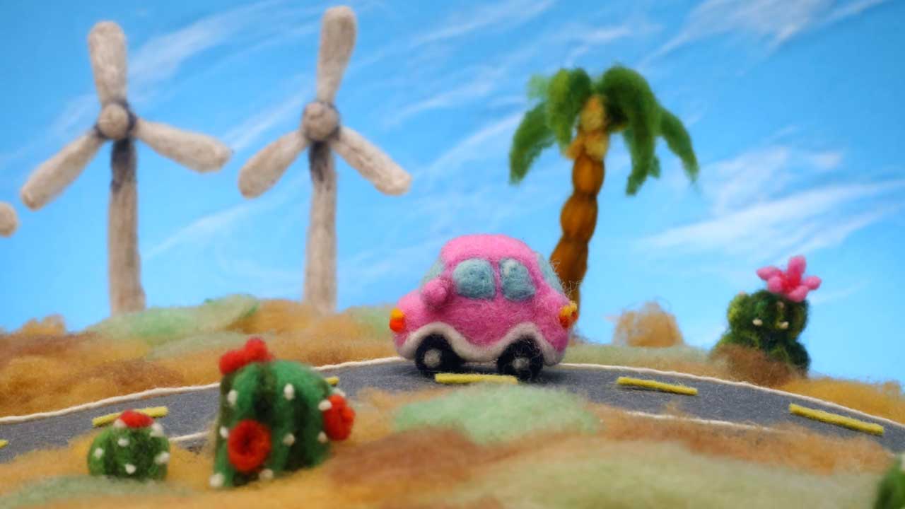 A felted pink car drives on a winding road past wind turbines, a palm tree, and cacti—all crafted from colorful felt—beneath a blue sky. Inspired by the Coachella 2026 visual identity film by Andrea Love and Hornet. Frame from Stash Magazine article.