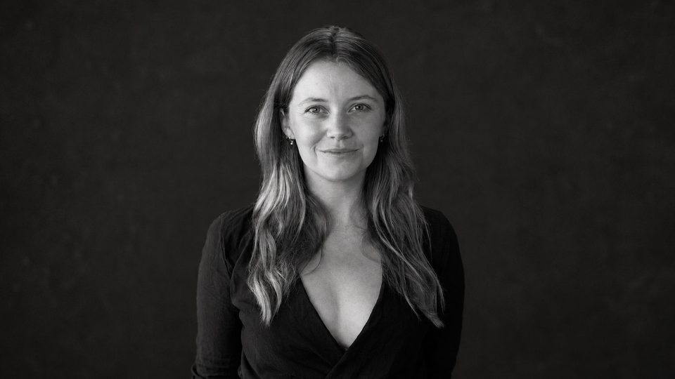 A black-and-white portrait of Coralie Tapper-Heckler with long hair, wearing a V-neck top against a dark background. Frame from Stash Magazine article.