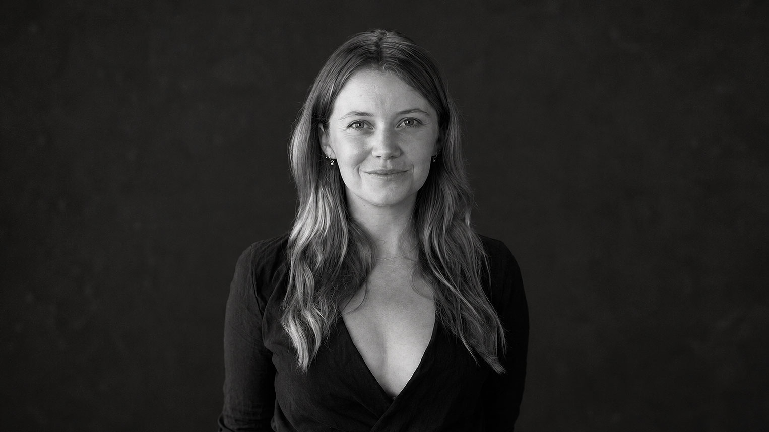 A black-and-white portrait of Coralie Tapper-Heckler with long hair, wearing a V-neck top against a dark background. Frame from Stash Magazine article.