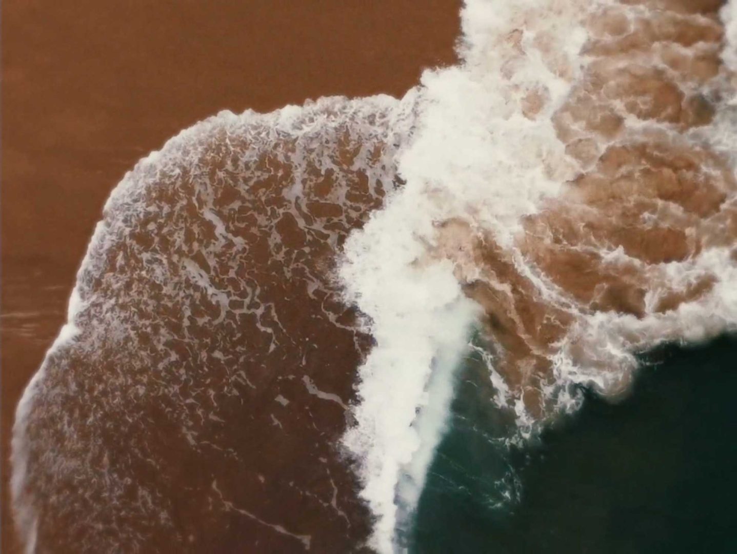 An aerial view of an ocean wave breaking onto a brown sandy beach, evoking the rhythmic textures of sonic studies. Frame from Stash Magazine article.