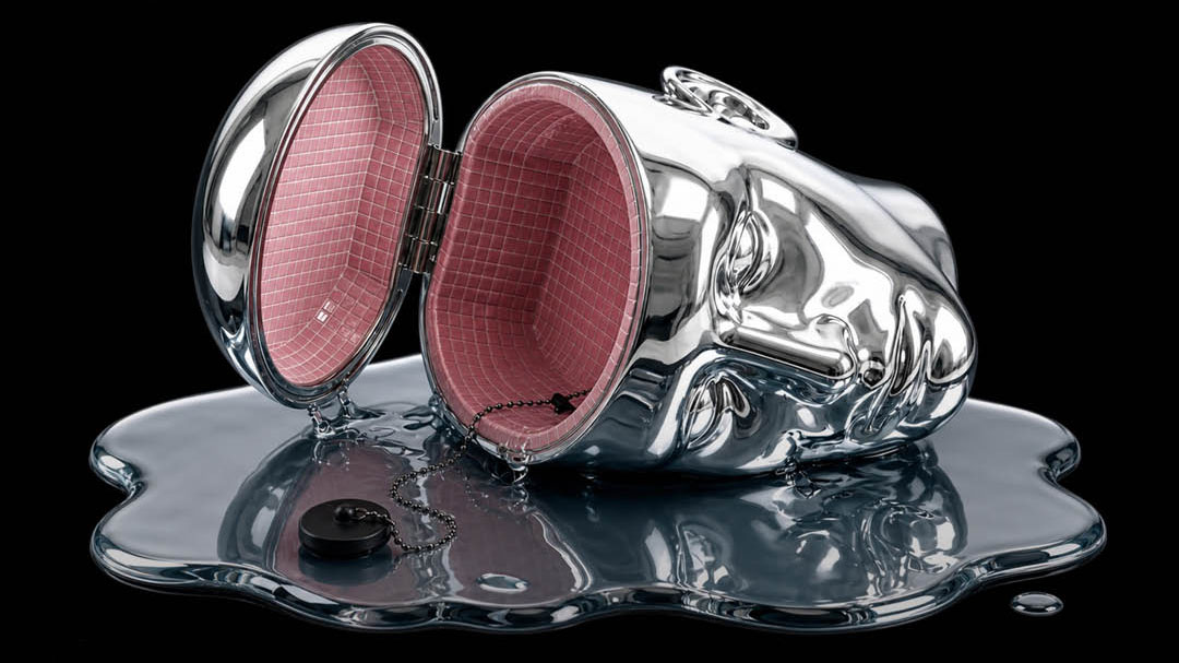 A shiny metallic purse shaped like a human face lies open, revealing a pink interior. This futuristic accessory, reminiscent of innovations spotlighted in the D&AD AI-REPORT-2026, rests on a puddle of melted metal with a detached button nearby, all set against a black background. Frame from Stash Magazine article.