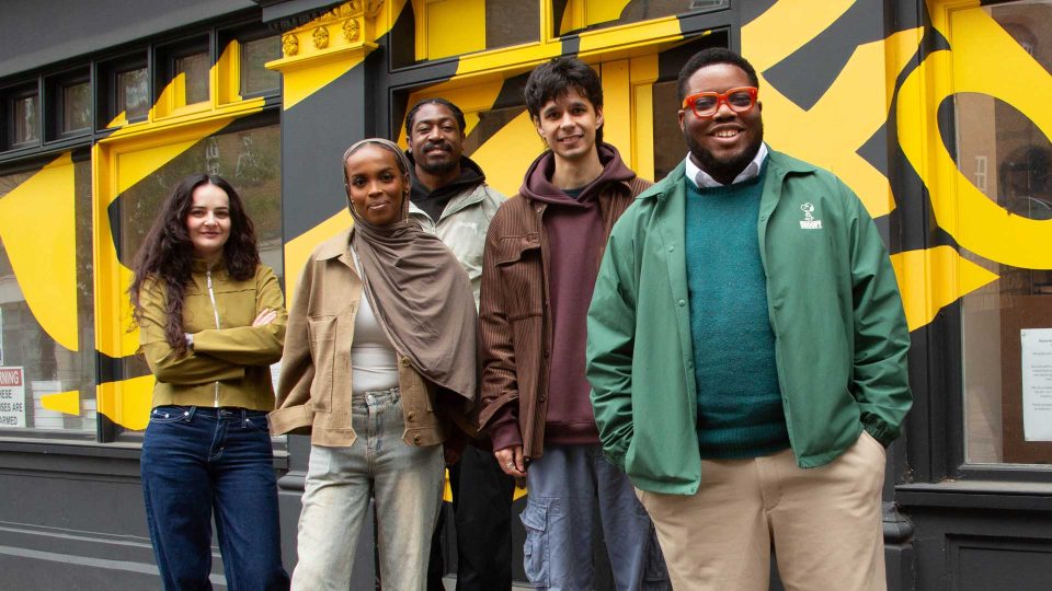 Five people stand smiling in front of a building with bold yellow and gray geometric designs. They wear casual clothing and appear relaxed, celebrating D&AD Shift at ten as they pose together as a group. Frame from Stash Magazine article.