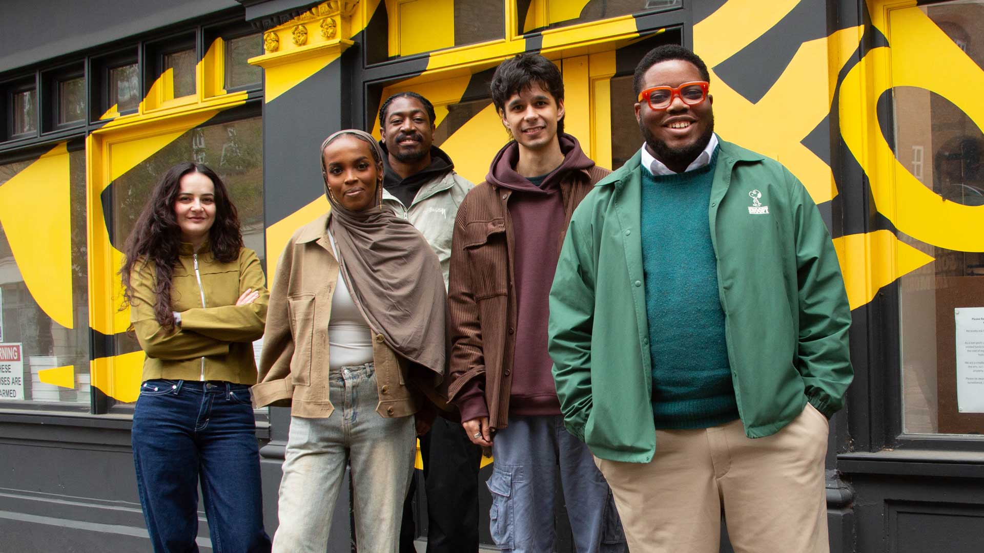 Five people stand smiling in front of a building with bold yellow and gray geometric designs. They wear casual clothing and appear relaxed, celebrating D&AD Shift at ten as they pose together as a group. Frame from Stash Magazine article.