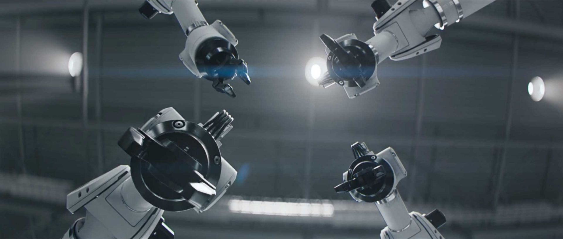 Four robotic arms with black grippers reach toward the center from above, set against an industrial ceiling with bright lights and a lens flare in the background—a striking visual reminiscent of the Dacia Robots commercial by Romain Chassaing. Frame from Stash Magazine article.