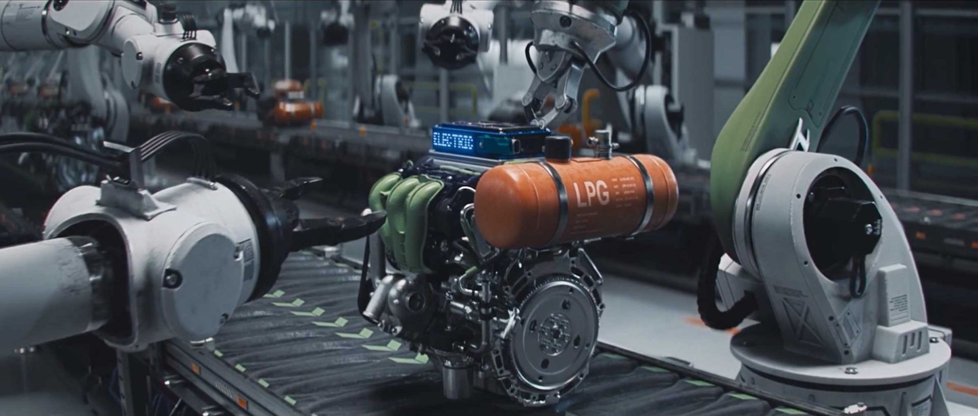 Robotic arms, reminiscent of the precision seen in the Dacia Robots commercial by Romain Chassaing, assemble an advanced engine with an orange LPG tank and green pipes on an automated conveyor in a modern factory setting. Frame from Stash Magazine article.