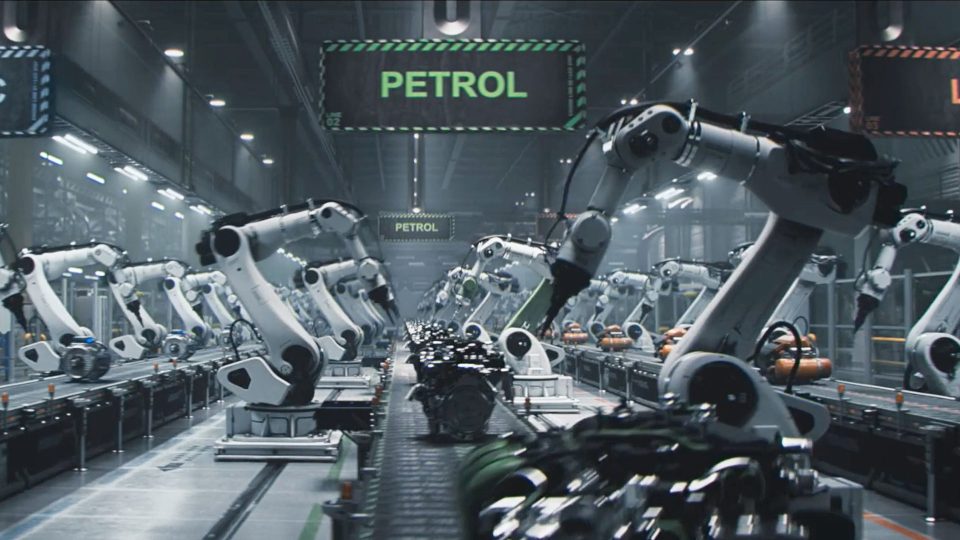 Rows of robotic arms assemble machinery on a conveyor belt in a modern factory, reminiscent of the precision seen in the Dacia Robots commercial by Romain Chassaing. A green sign labeled PETROL hangs overhead, indicating the production focus. The setting is industrial and high-tech. Frame from Stash Magazine article.