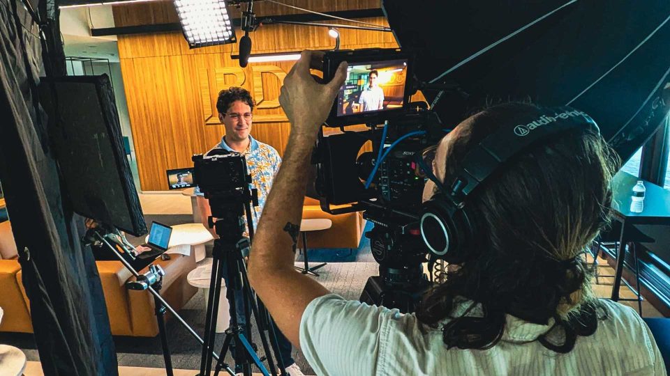 A videographer filming a man during an interview as part of Demo Duck’s long-form documentary videos services in a modern office. Frame from Stash Magazine article.