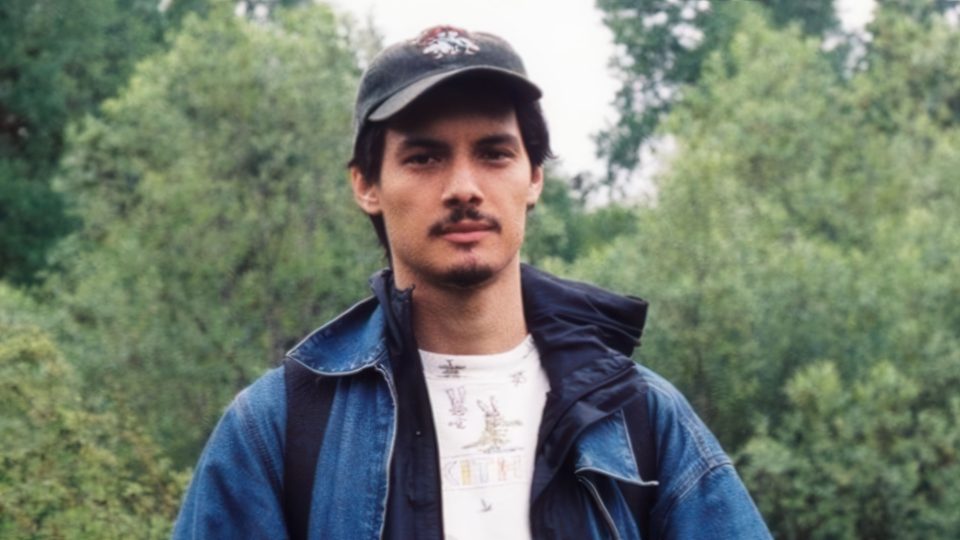 A man wearing a black cap, blue jacket, and white t-shirt stands outdoors with green trees in the background, capturing the confident presence reminiscent of Director Liam Archibald Signs to Blinkink Roster. Frame from Stash Magazine article.