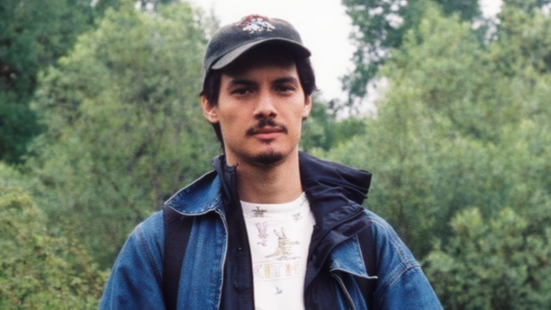A man wearing a black cap, blue jacket, and white t-shirt stands outdoors with green trees in the background, capturing the confident presence reminiscent of Director Liam Archibald Signs to Blinkink Roster. Frame from Stash Magazine article.