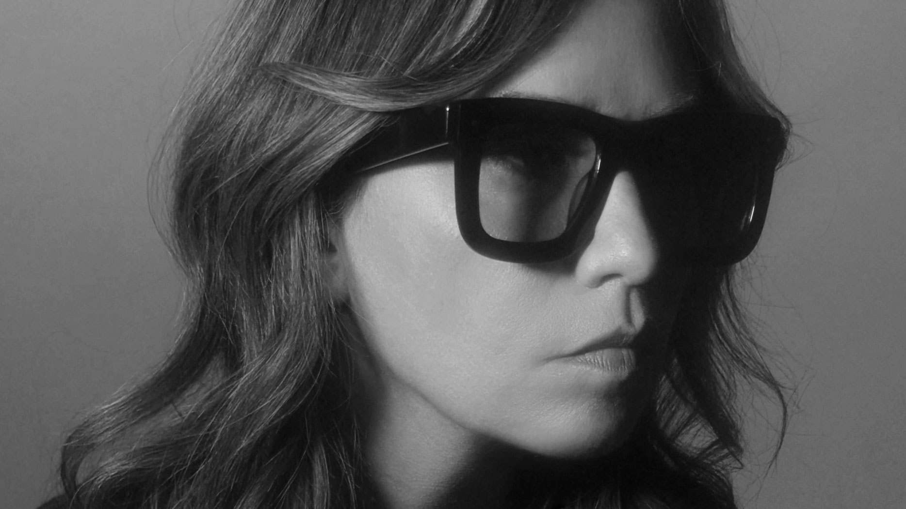 A person with wavy hair wearing large, dark sunglasses looks serious in this black-and-white close-up portrait by director Sarah McColgan. Frame from Stash Magazine article.