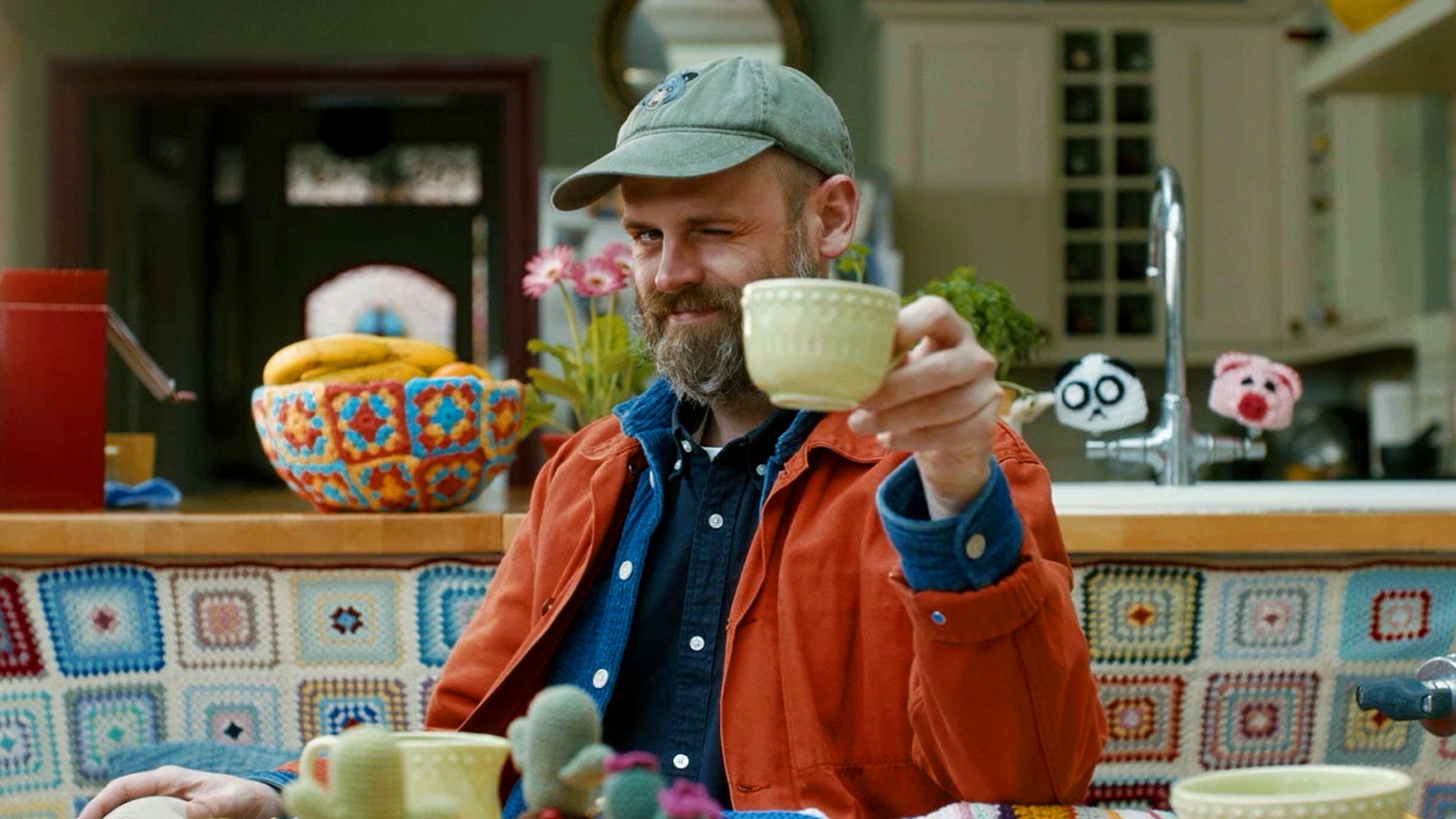 Director Zac Ella winks while holding up a yellow mug in a kitchen with crocheted decor. Frame from Stash Magazine article.
