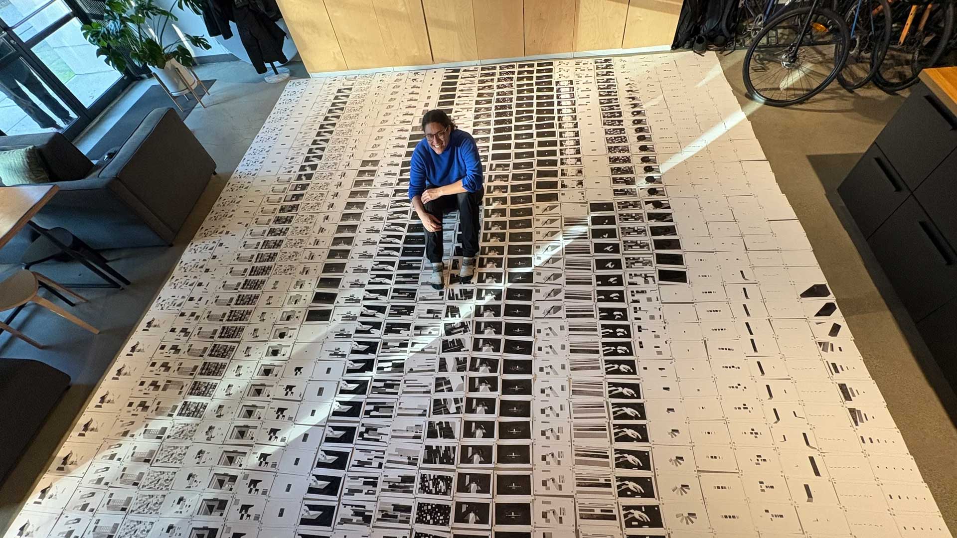 A person in a blue sweater sits in the center of a room on a large grid of printed photo contact sheets, evoking a sense of distance among the Ordinary Folk. Frame from Stash Magazine article.