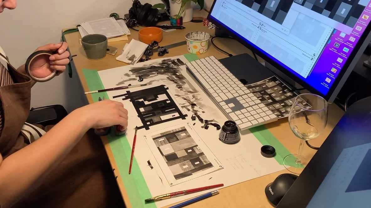 A person at a desk uses a brush and black ink to draw architectural floor plans on paper, reflecting the artistic process seen in the Ordinary Folk short film Distance, while a monitor shows digital design software in the background. Frame from Stash Magazine article.