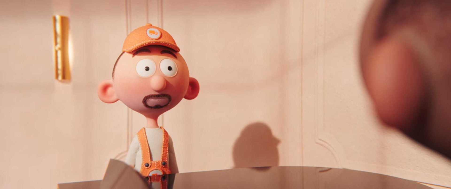 A claymation-style character with a mustache and beard stubble, wearing an orange cap and overalls, stands behind a desk looking surprised; in this Duet short film by Léo Brunel, another blurred figure is partially visible in the foreground. Frame from Stash Magazine article.