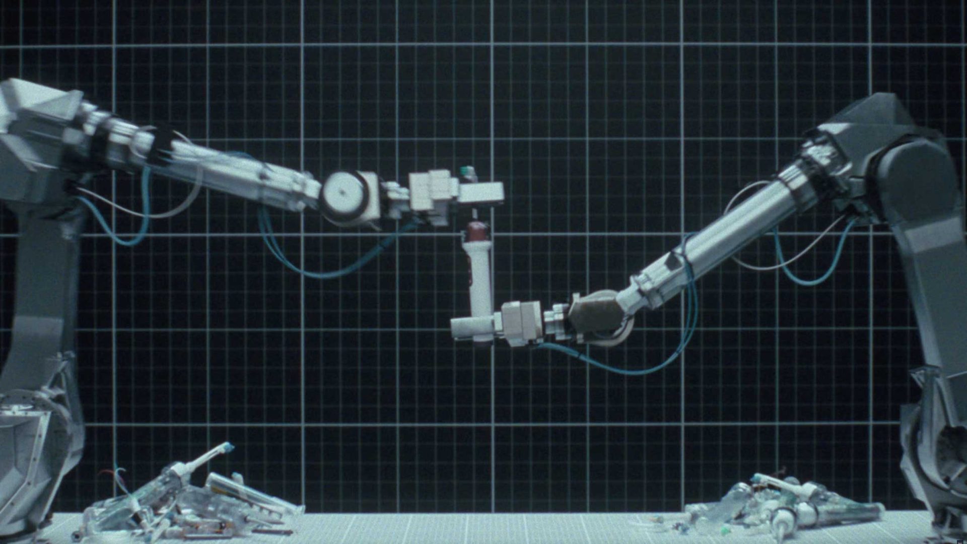 Two robotic arms face each other over a grid-lined background, holding a small Dyson CameraJet part between them. Disassembled robot parts are scattered on the surface below, creating a scene reminiscent of Will Dohrn’s innovative designs. Frame from Stash Magazine article.