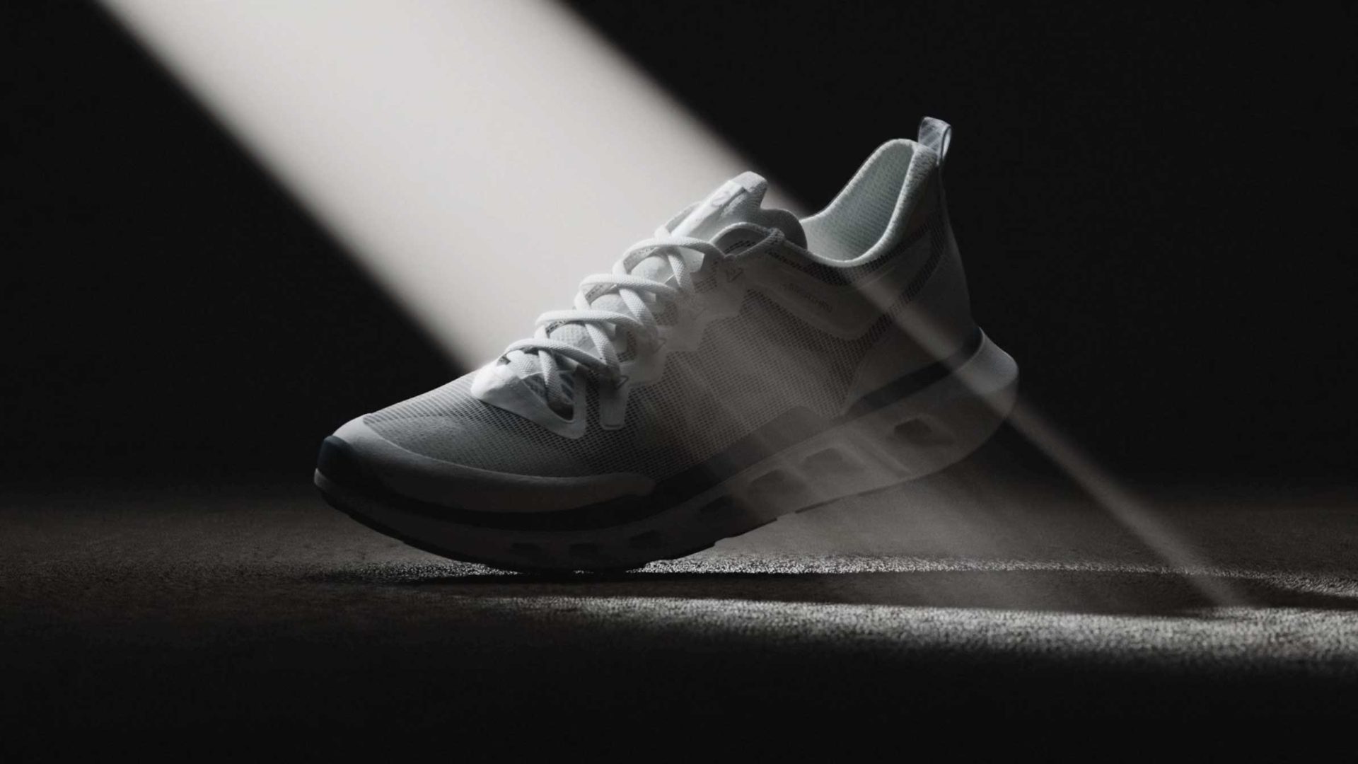 A white ECCO Biom 720 athletic sneaker is illuminated by dramatic beams of light in a dark product film setting, highlighting its details and mesh texture as it rests on a textured surface. Frame from Stash Magazine article.