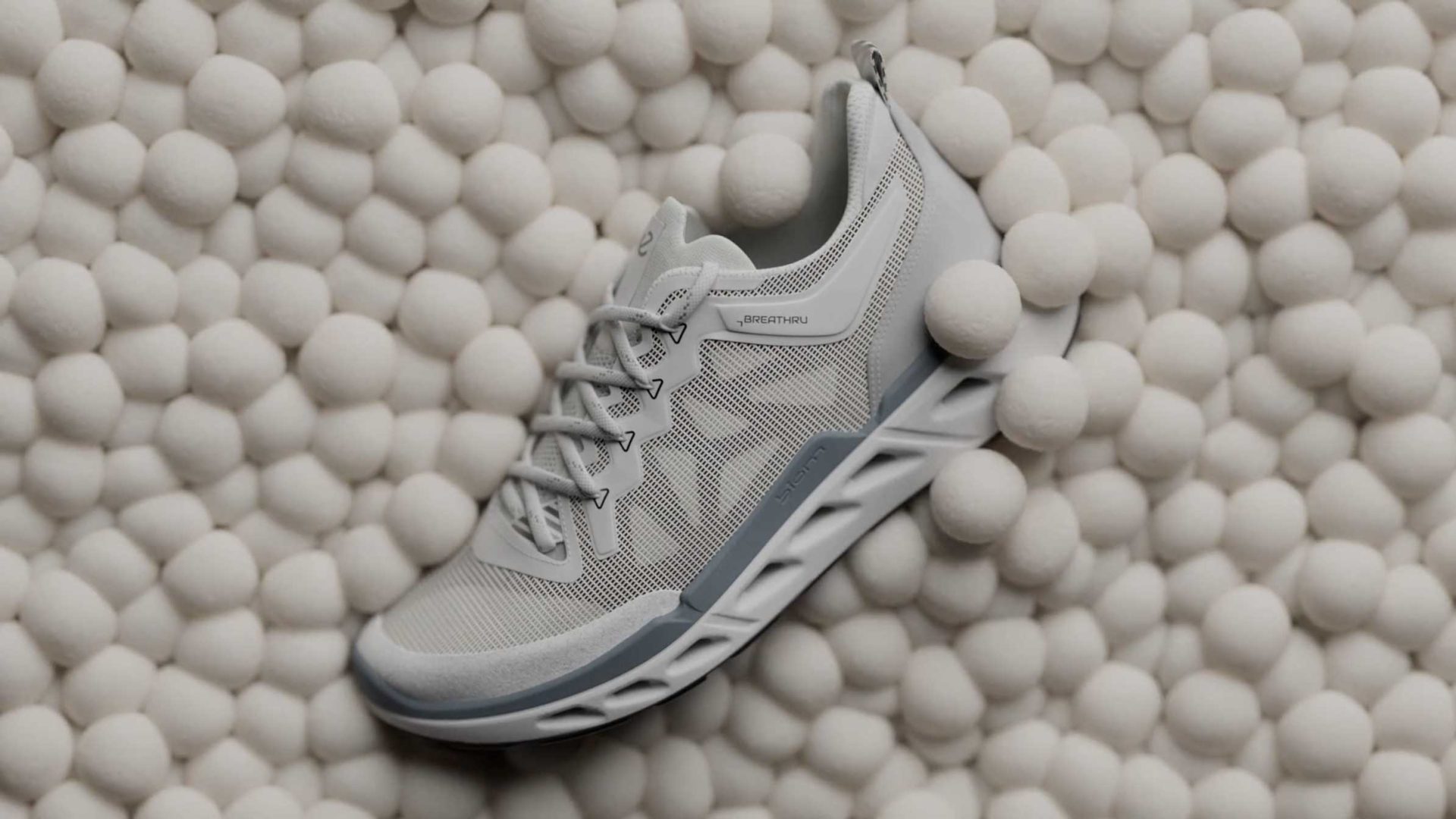 A light gray ECCO Biom 720 athletic sneaker with white and gray accents rests on a surface covered with numerous small, soft white balls. The shoe has a textured sole and mesh upper, showcasing its modern and sporty look in this product film. Frame from Stash Magazine article.