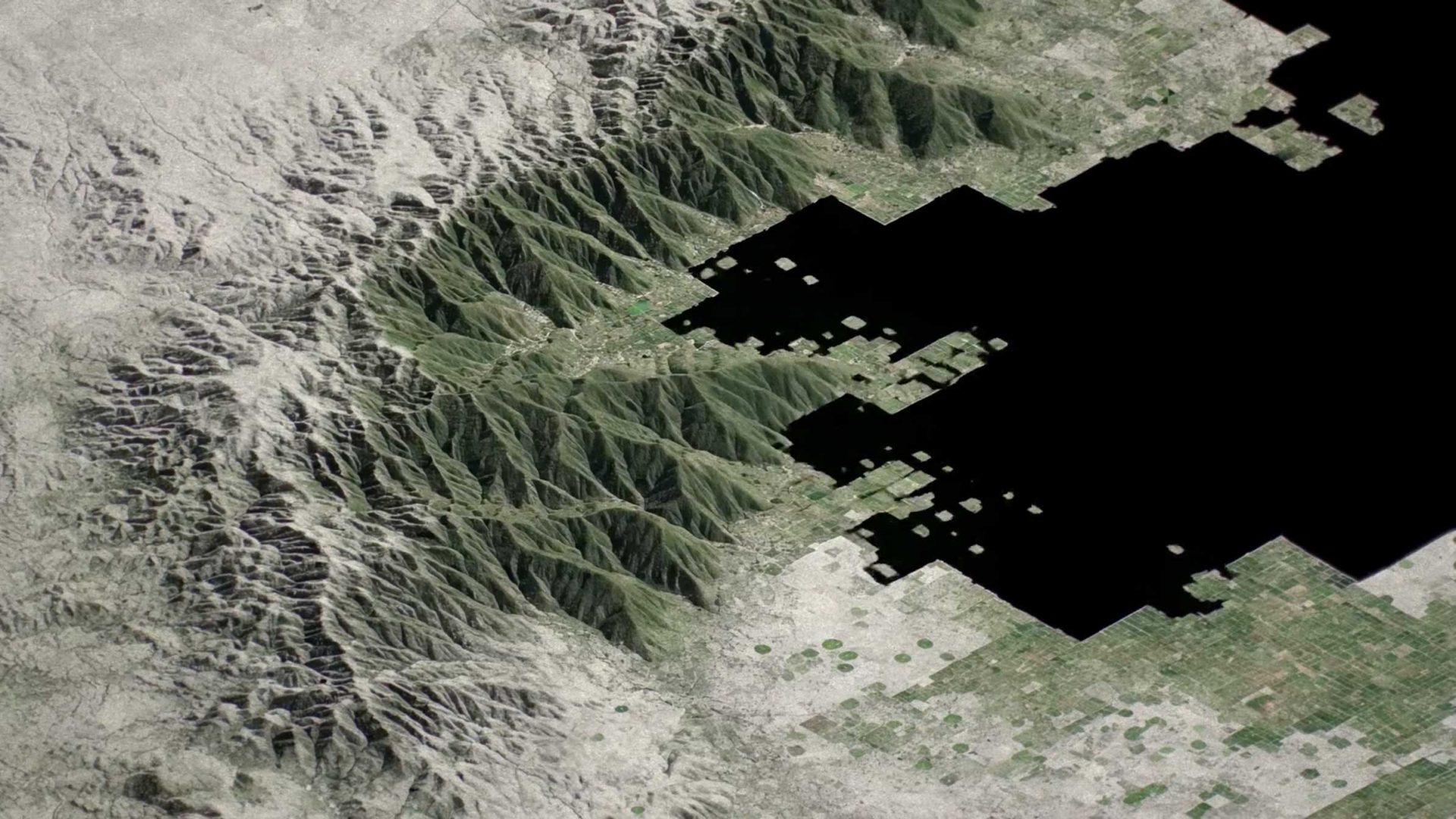 Satellite view of a mountain range bordering a flat, gridded landscape, partially obscured by Ege Carpets geometric cutouts. Frame from Stash Magazine article.