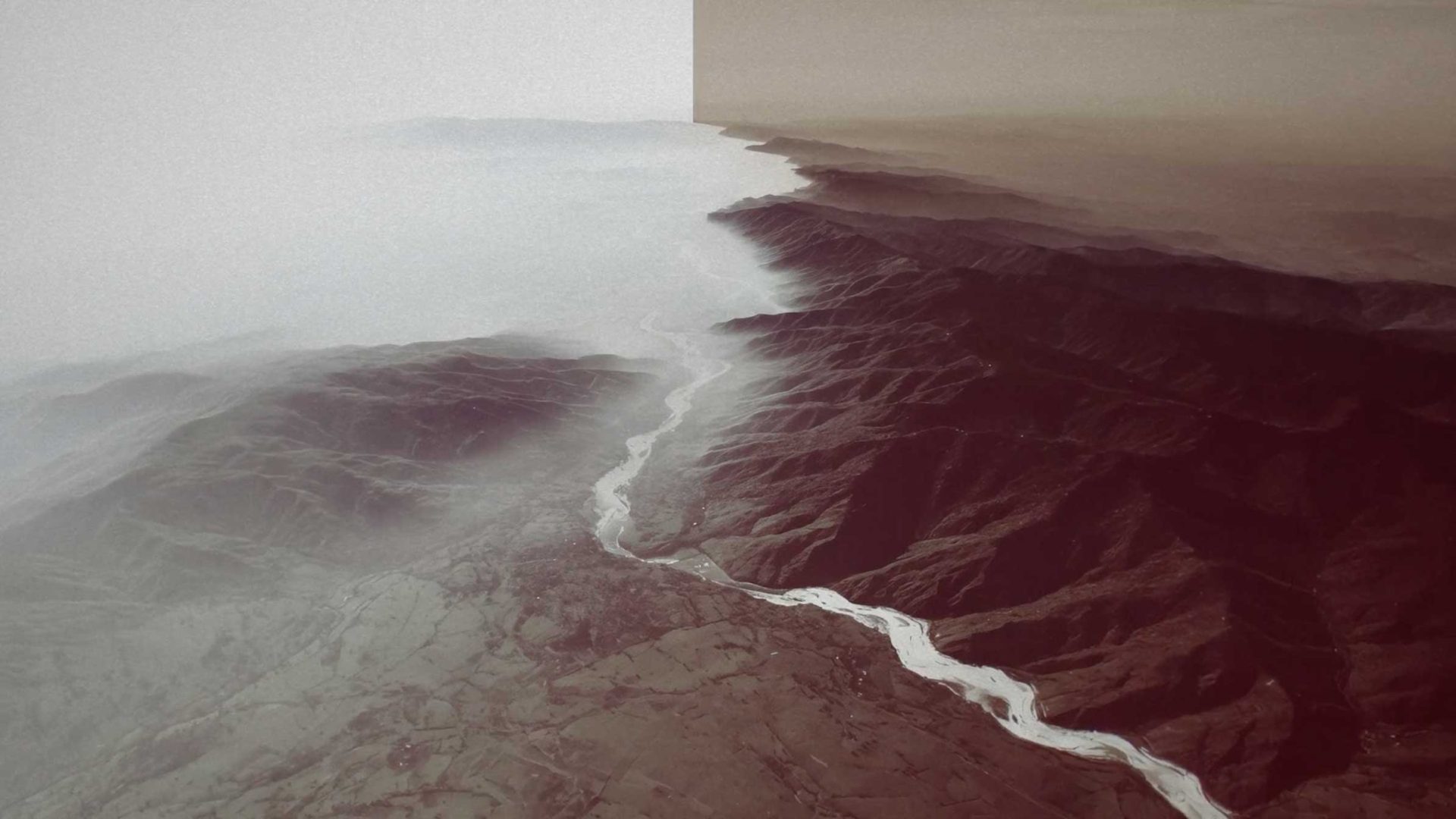 An aerial view of a river from a Gensler x Ege Carpets film, winding through mountainous terrain split between a hazy, white-toned landscape and a sepia-toned landscape. Frame from Stash Magazine article.