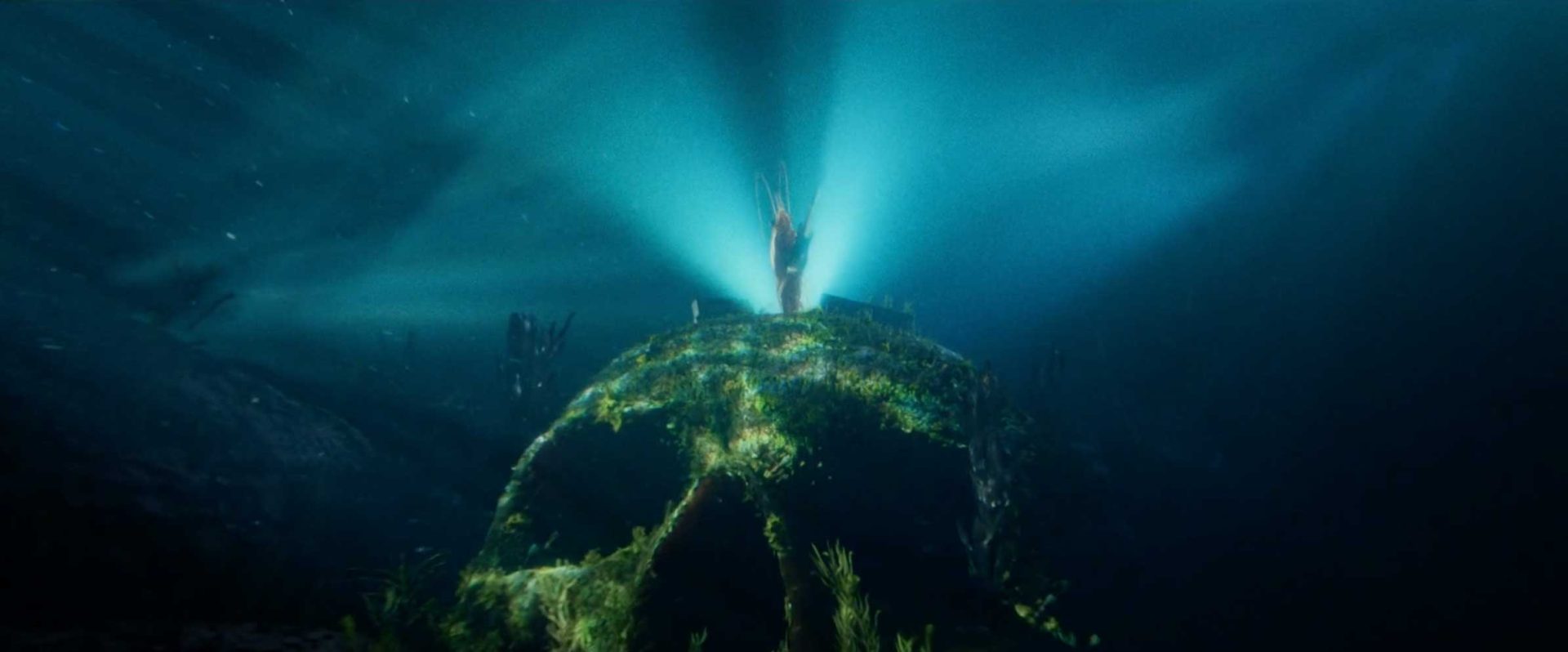 A glowing figure stands atop a large, mossy skull-shaped rock underwater on Skull Island, illuminated by blue light rays and surrounded by aquatic plants—an ethereal guardian amid the mysteries where only the bravest dare to Eat the Wild. Frame from Stash Magazine article.