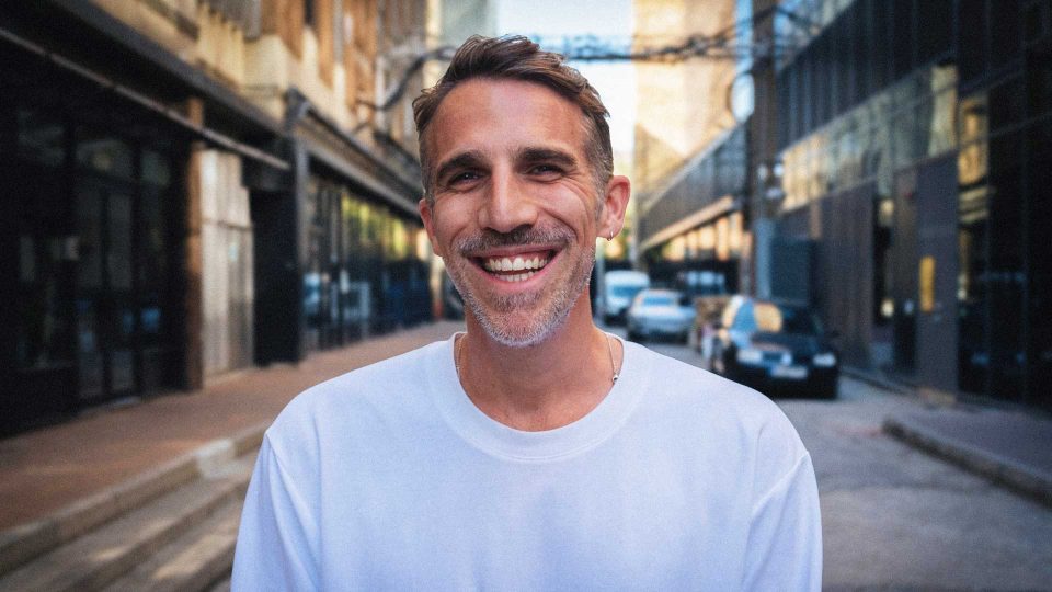 A man with short brown hair and a beard, wearing a white shirt, smiles while standing on a city street with buildings and cars in the background—this is Ed Christie, Lead Motion Designer at Electric Theatre Collective. Frame from Stash Magazine article.