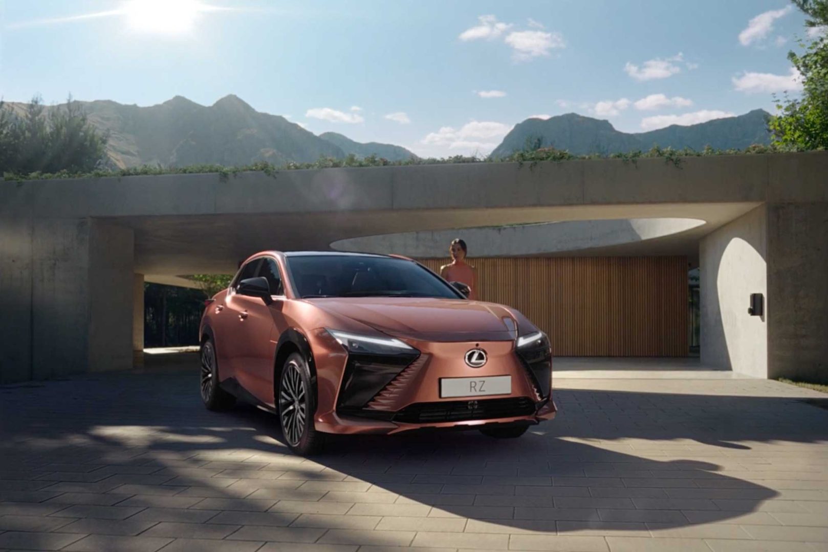A bronze Lexus RZ electric SUV is parked outside a modern house with curved concrete architecture; mountains and a blue sky with clouds are in the background, and a person—perhaps from the Electrical Theatre Collective—stands near the car. Frame from Stash Magazine article.