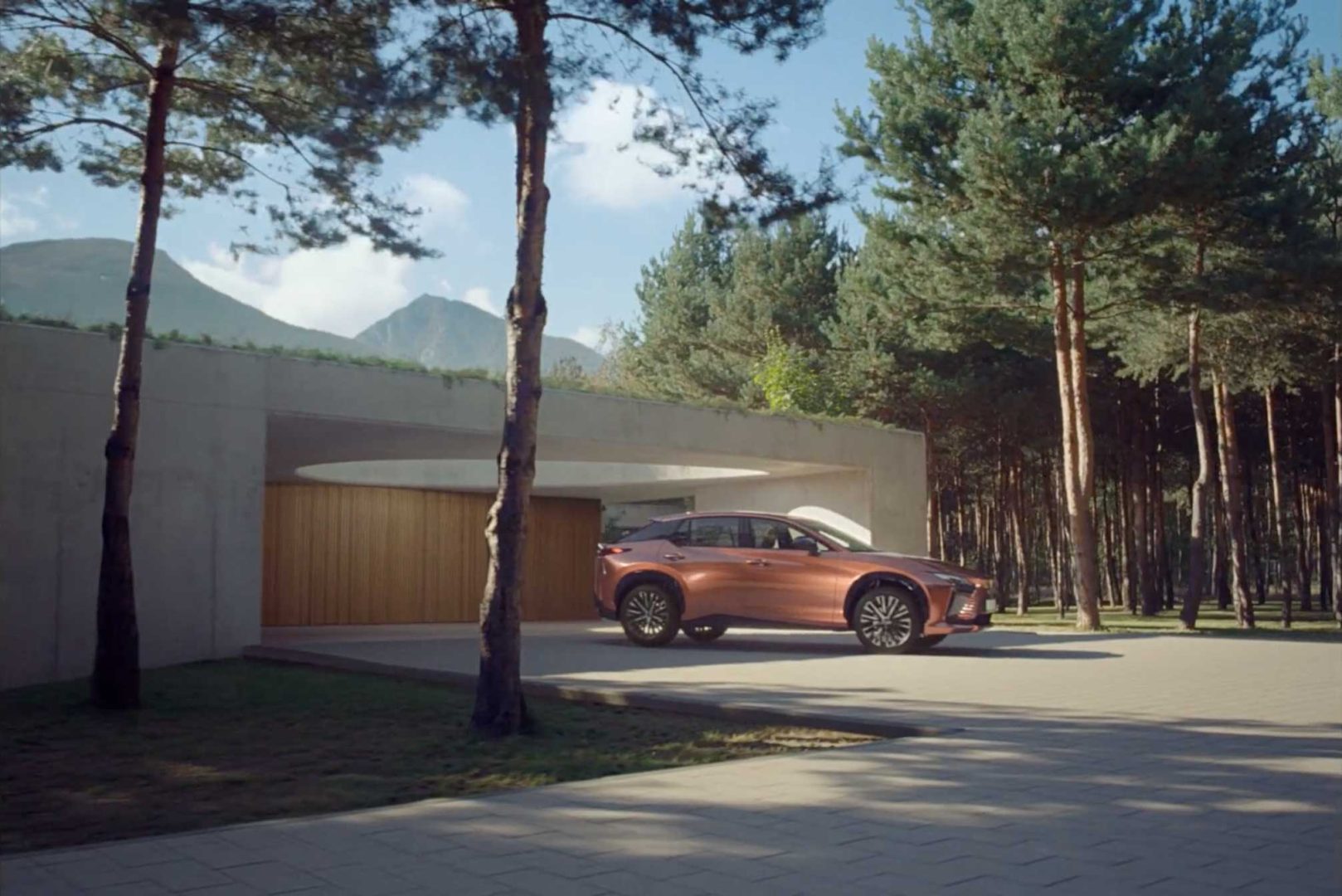 A bronze SUV, possibly the Lexus RZ, is parked in front of a modern house with a wooden garage—Electrical Theatre Collective digs into the "Details" as tall pine trees and mountains complete the scene under a partly cloudy sky. Frame from Stash Magazine article.