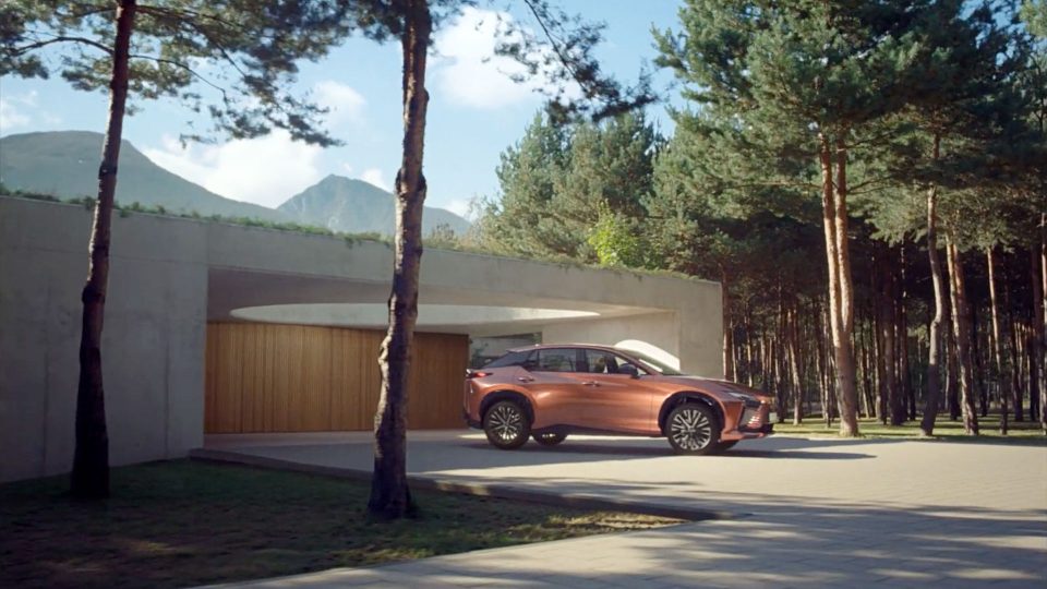 A bronze SUV is parked in front of a modern house with wooden garage doors, as the Electrical Theatre Collective digs into the "Details" of Lexus RZ, surrounded by tall pine trees and mountains on a sunny day. Frame from Stash Magazine article.