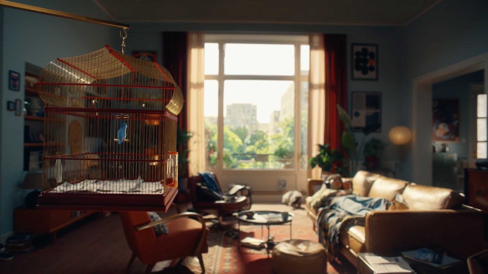 A blue parakeet sits in a hanging birdcage in a cozy, sunlit living room—by the large window, tan sofa, and scattered books, an Erste Bank leaflet rests among the plants and blankets. Frame from Stash Magazine article.