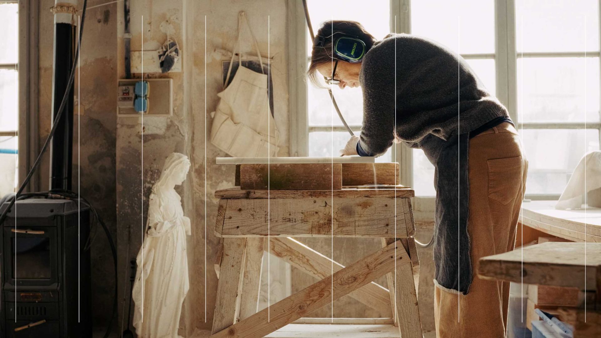 A person wearing earmuffs uses a power tool to carve an Exemplar typeface into a piece of stone on a wooden workbench in a studio. Frame from Stash Magazine article.