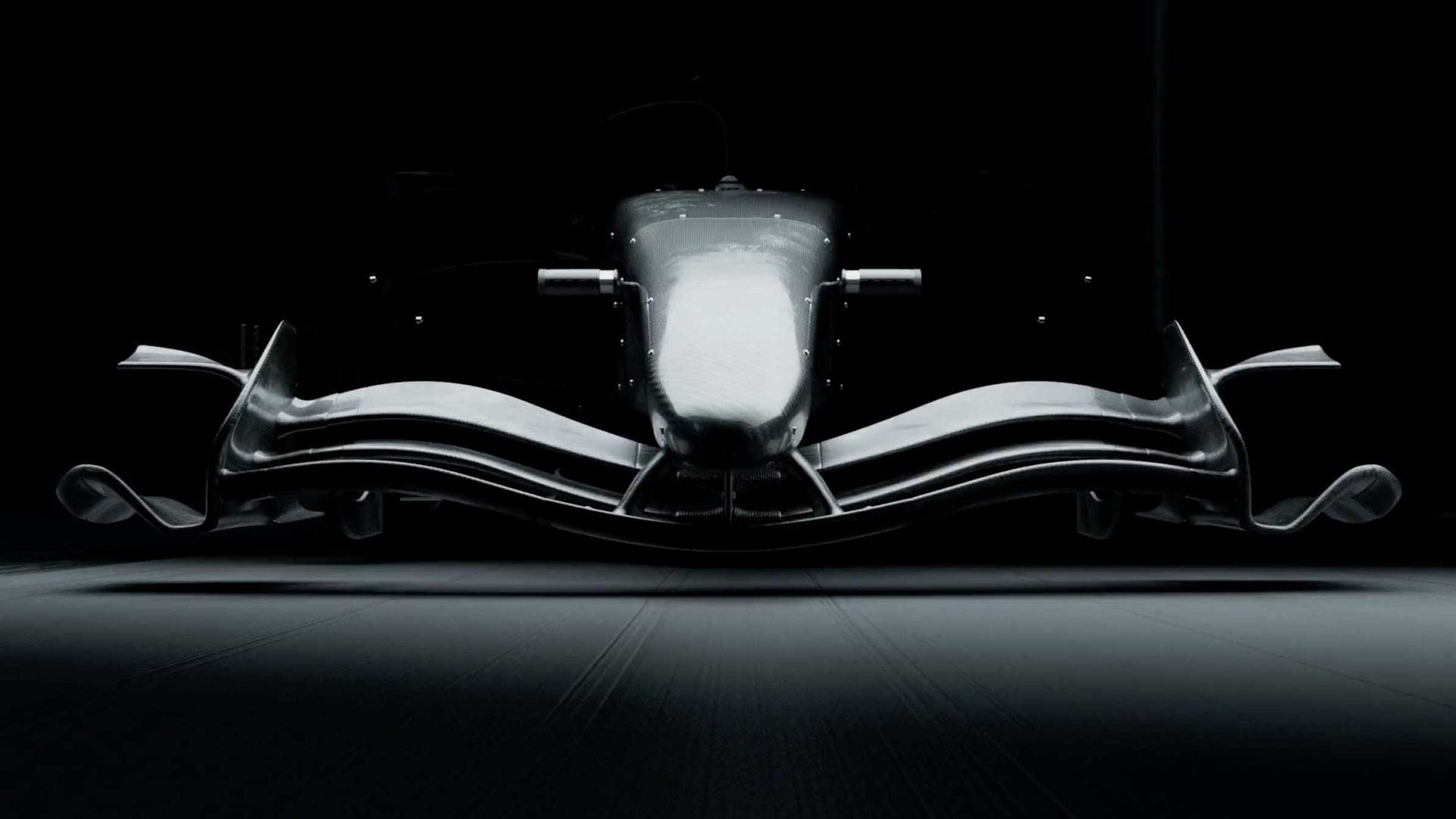 A dramatic, low-angle front view of a modern Formula 1 car from the F1 2026 Opening Titles Process Reel by DissEmbargo, mostly in shadow, highlighting its aerodynamic nose, front wing, and sleek curves against a dark background. Frame from Stash Magazine article.