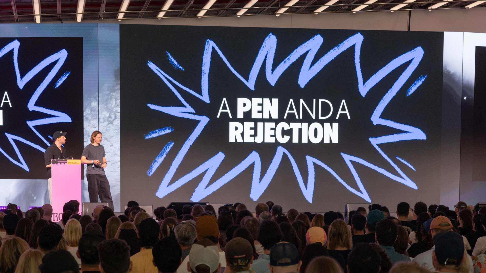 Two speakers stand onstage in front of a large screen displaying the words A PEN AND A REJECTION, with a blue starburst design, as a seated audience watches—a scene reminiscent of a Stash Interview with Dirk Schuster from FOREAL. Frame from Stash Magazine article.
