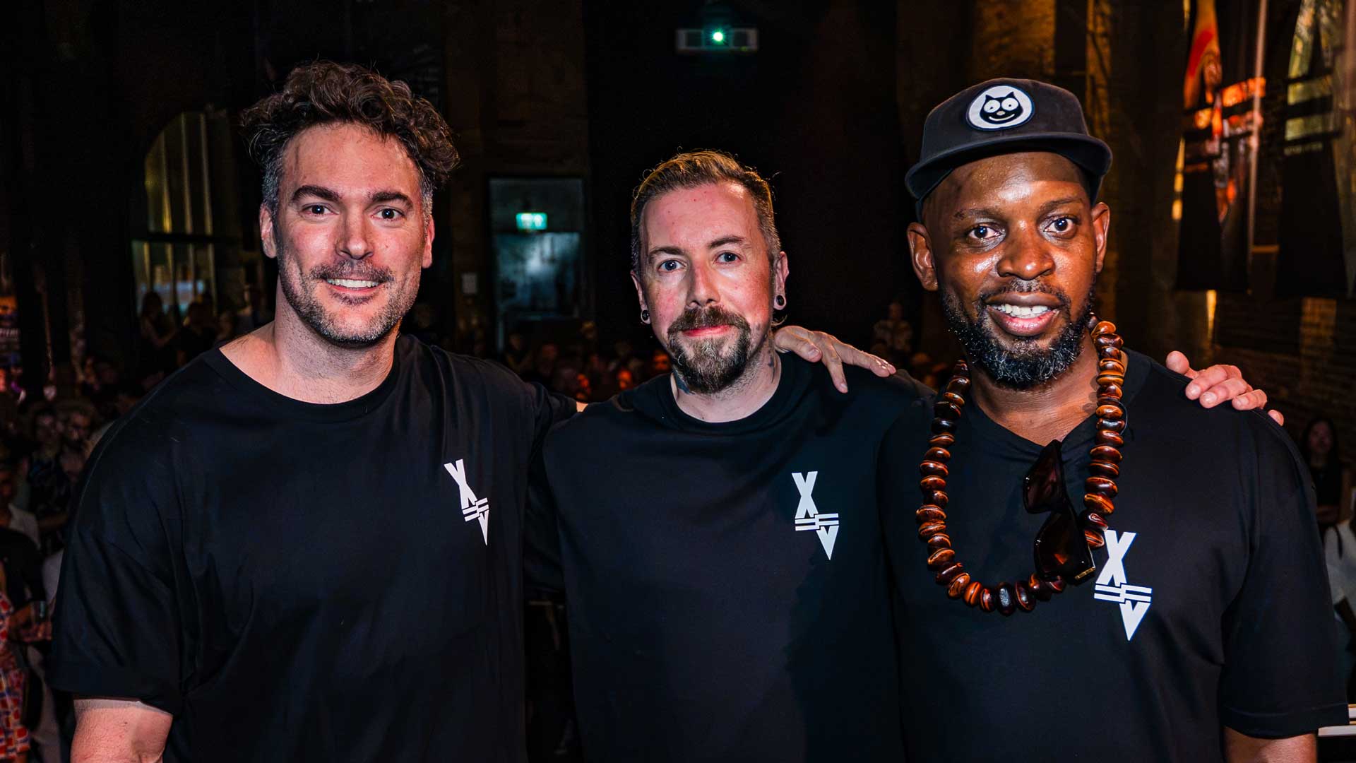 Three men, including Factory-Fifteen's Kev McCrae, wear matching black shirts and smile at the camera in a dimly lit indoor setting. One sports a beaded necklace. An audience and banners can be seen in the background. Frame from Stash Magazine article.