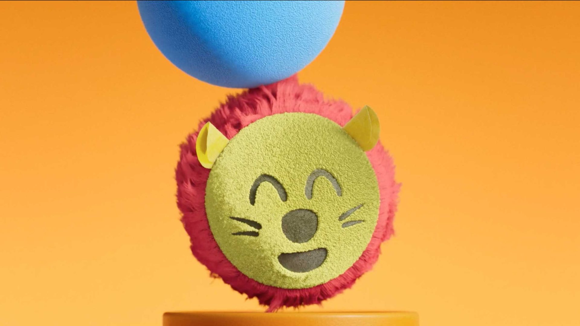 A round, plush lion face with a green snout, yellow ears, and fuzzy red mane is shown against an orange background. A blue ball from Fat Brain Toys sits above the lion’s head. Frame from Stash Magazine article.