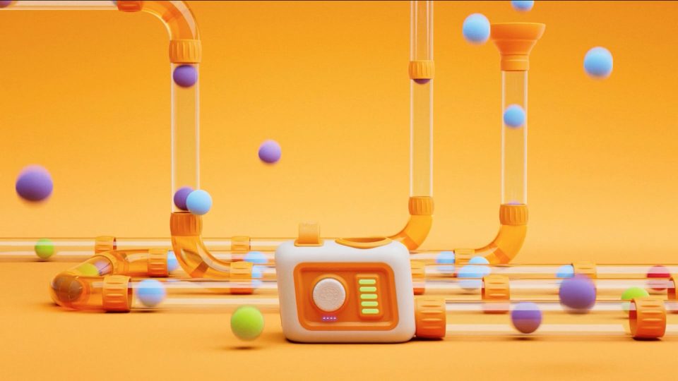 A white and orange device with glowing indicators sits among clear tubes, while colorful balls—featured in the Fat Brain Toys Air Toobz Film by Frame48—float and travel through the tubes, all set against an orange background. Frame from Stash Magazine article.