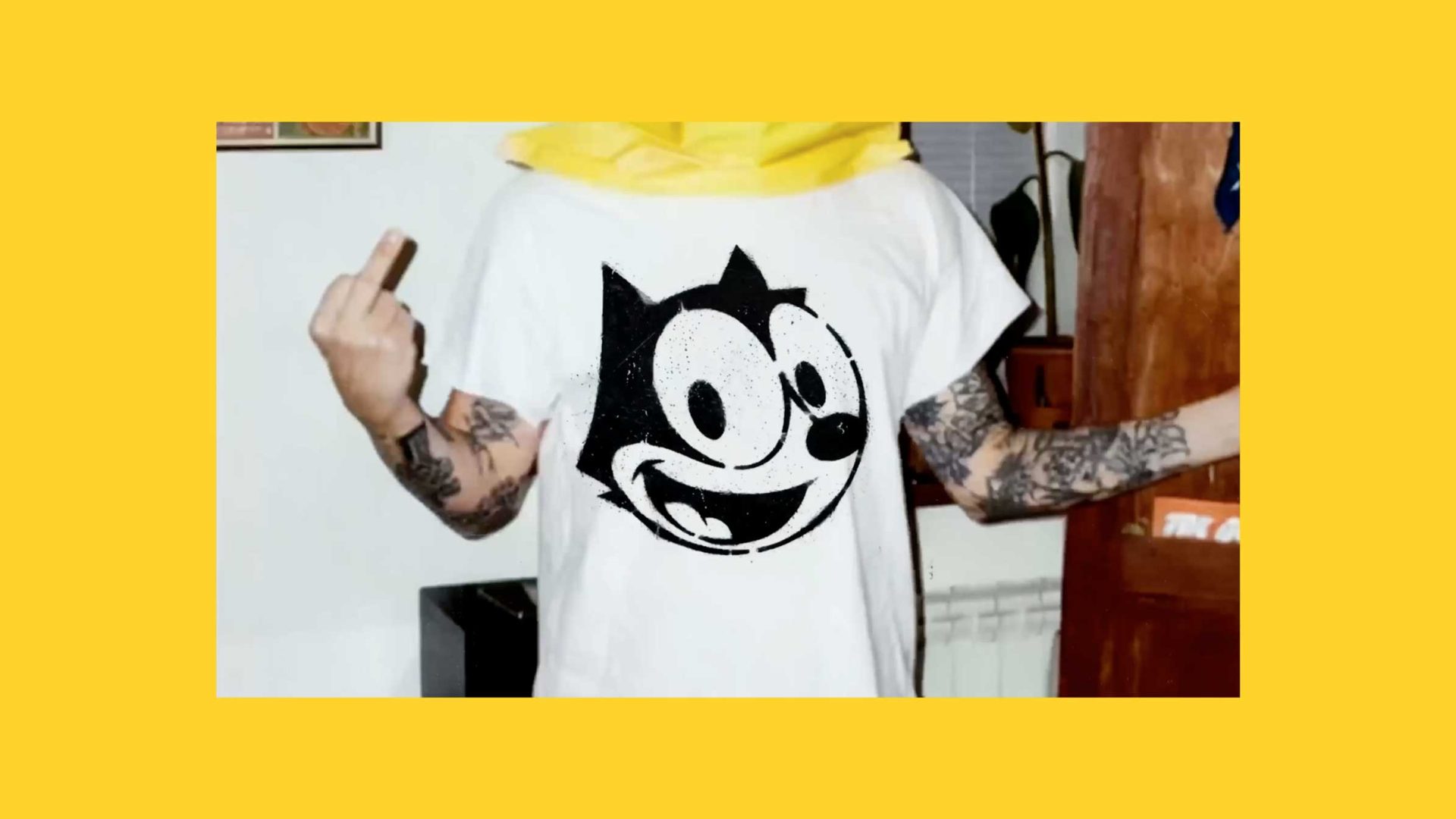 A person with tattooed arms wears a white T-shirt featuring a Felix the Cat cartoon face and stands indoors, raising their left middle finger. The image, perfect for a Soda Creative sizzle reel, has a yellow border. Frame from Stash Magazine article.