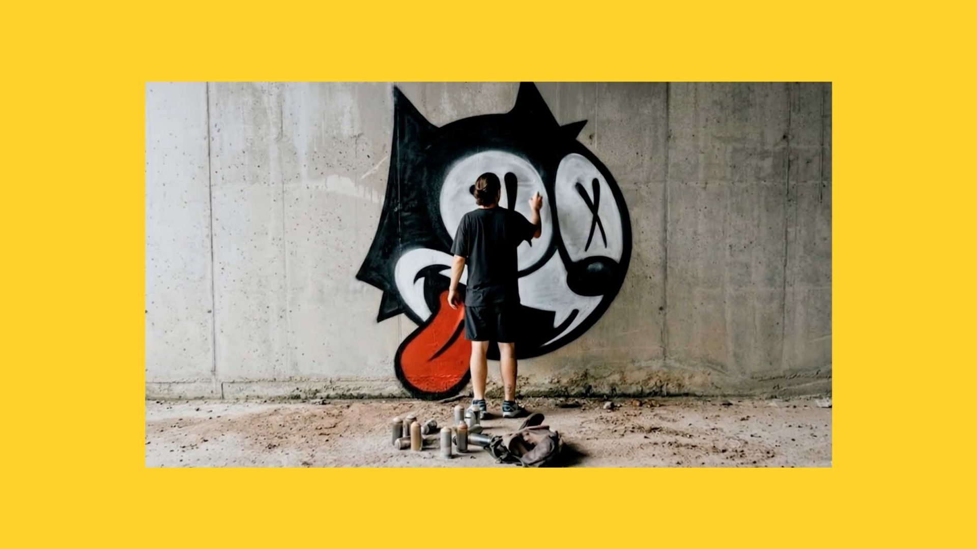 A person spray paints a cartoonish black-and-white Felix the Cat with a red tongue on a concrete wall. Several spray cans and a backpack are on the ground, all framed by a yellow border in this Soda Creative sizzle reel moment. Frame from Stash Magazine article.