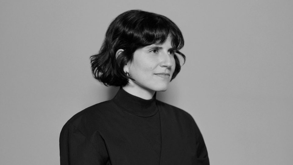 A woman with short, dark hair wearing a high-collared, dark top looks slightly to the right and smiles softly. The image of director Andrea Devia-Nuño is in black and white against a plain, light background, evoking a Final Frontier spirit. Frame from Stash Magazine article.