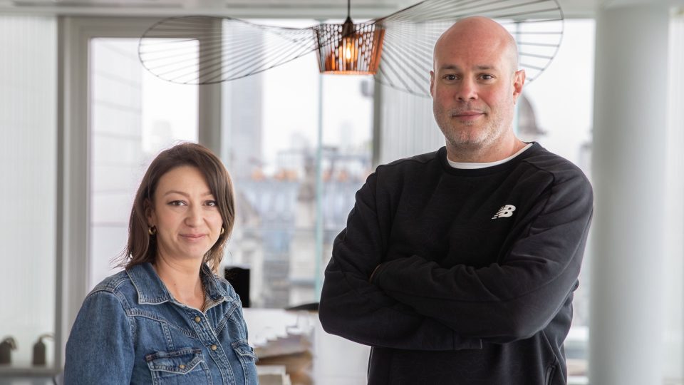 A woman in a denim jacket and a man in a black sweatshirt stand together in a bright modern office with large windows and a city view—Framestore VFX Supervisor Dan Lorenzini and EP Kayleigh Dugdale. Frame from Stash Magazine article.