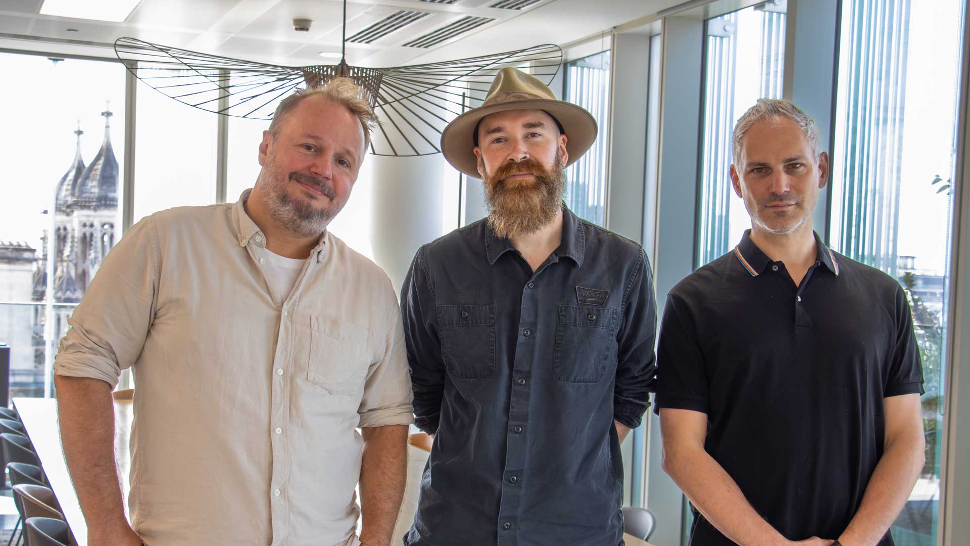 Three men—Sean-Cooper, Ric-Comline, and Thierry-Levy—stand indoors by large windows, smiling and facing the camera. One wears a hat and has a beard, while the others are clean-shaven. The modern office setting features city buildings visible outside. Frame from Stash Magazine article.