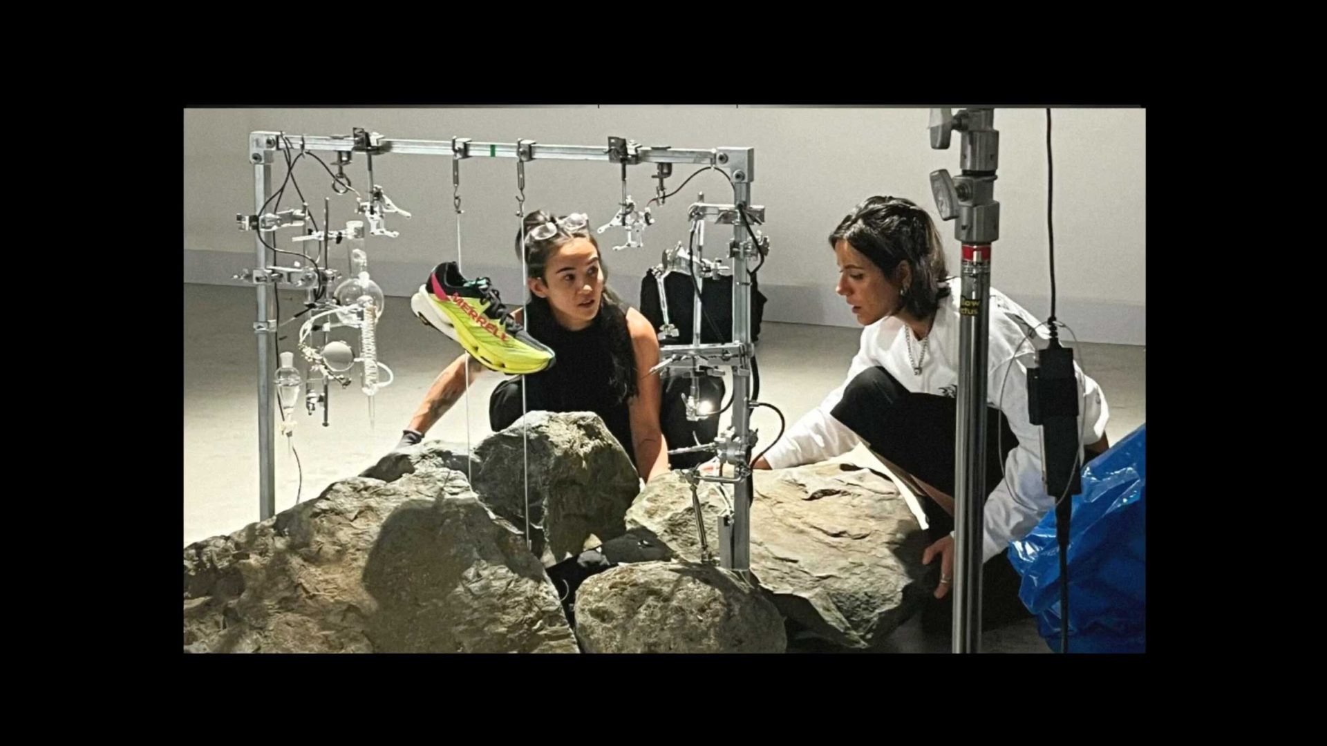 Two women kneel by large rocks, adjusting a complex metal rig holding a single Merrell running shoe. The setup, part of a sleek FutureDeluxe campaign for the MTL SpeedARC Peak, is in an indoor studio space with professional lighting. Frame from Stash Magazine article.