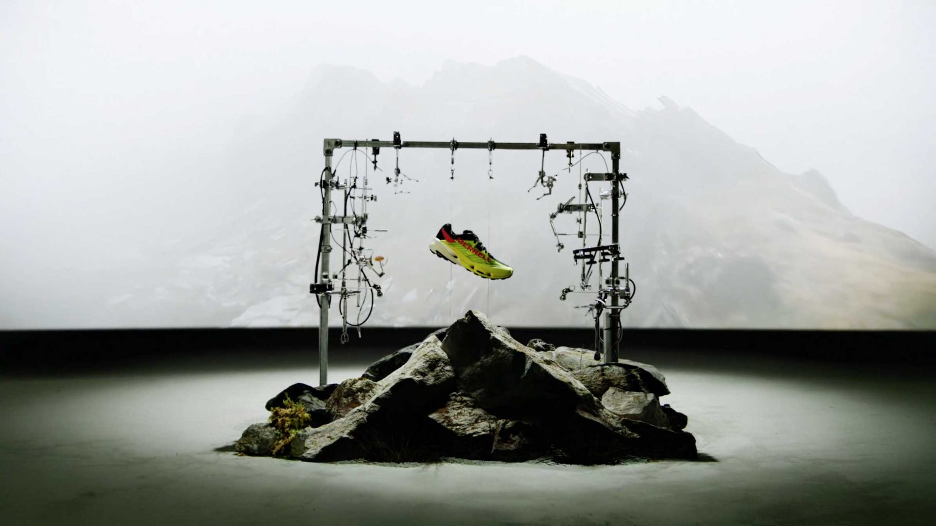 A bright Merrell running shoe, the MTL SpeedARC Peak, is suspended mid-air by wires above a rocky mound, with a misty mountain in the background, creating a dramatic and futuristic display. Frame from Stash Magazine article.