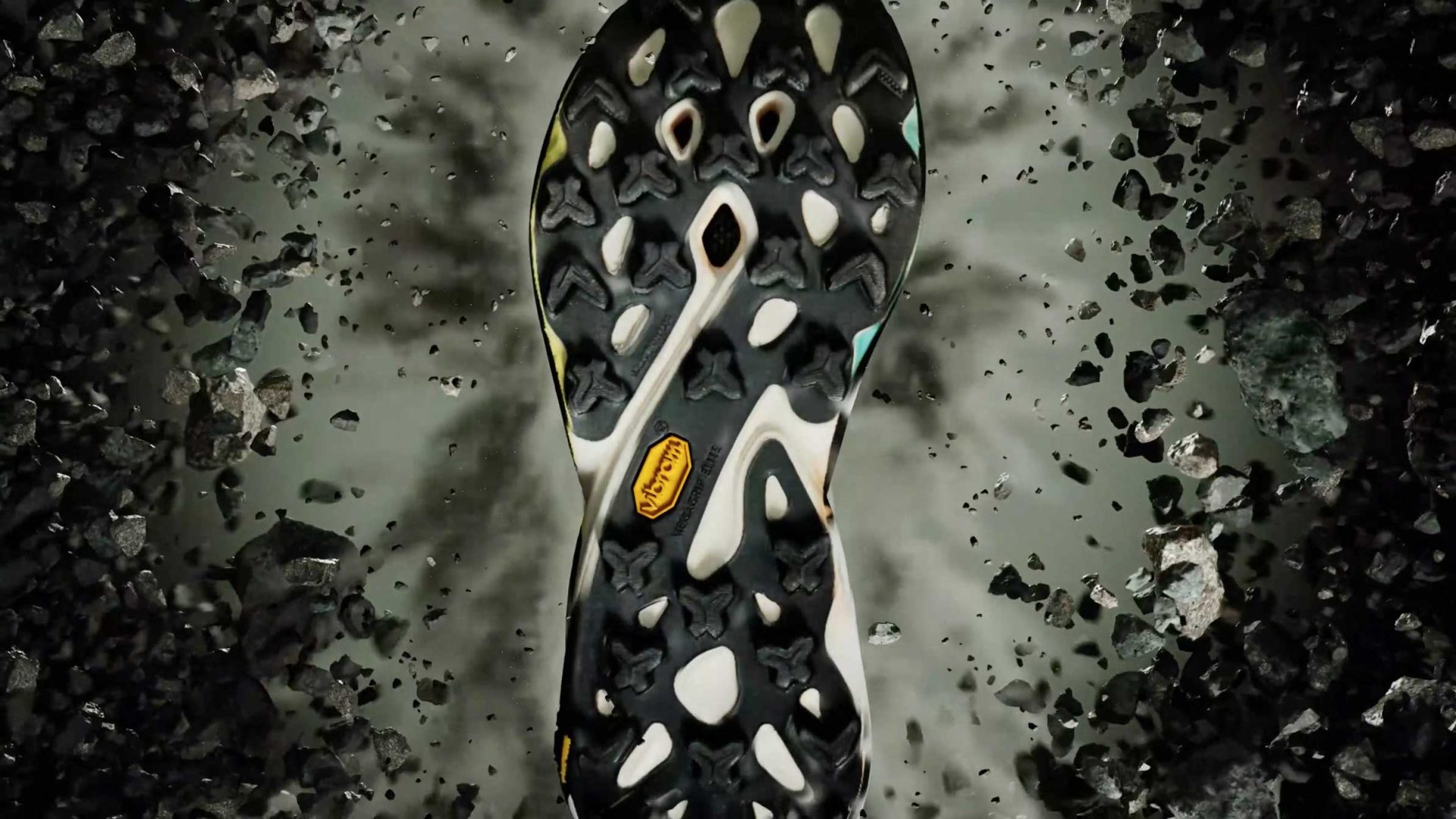 Close-up of a black and white patterned shoe sole, designed by Merrell for the MTL SpeedARC Peak, with debris and rocks flying around it—emphasizing the rugged tread and advanced outdoor functionality captured in FutureDeluxe’s signature visual style. Frame from Stash Magazine article.