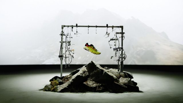 A colorful Merrell sneaker is suspended by wires in a metal frame above rocky terrain, with a misty mountainous backdrop, evoking the cutting-edge design of the MTL SpeedARC Peak. Frame from Stash Magazine article.