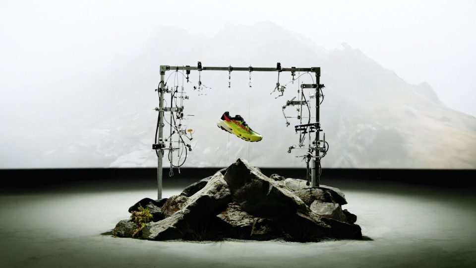 A colorful Merrell sneaker is suspended by wires in a metal frame above rocky terrain, with a misty mountainous backdrop, evoking the cutting-edge design of the MTL SpeedARC Peak. Frame from Stash Magazine article.
