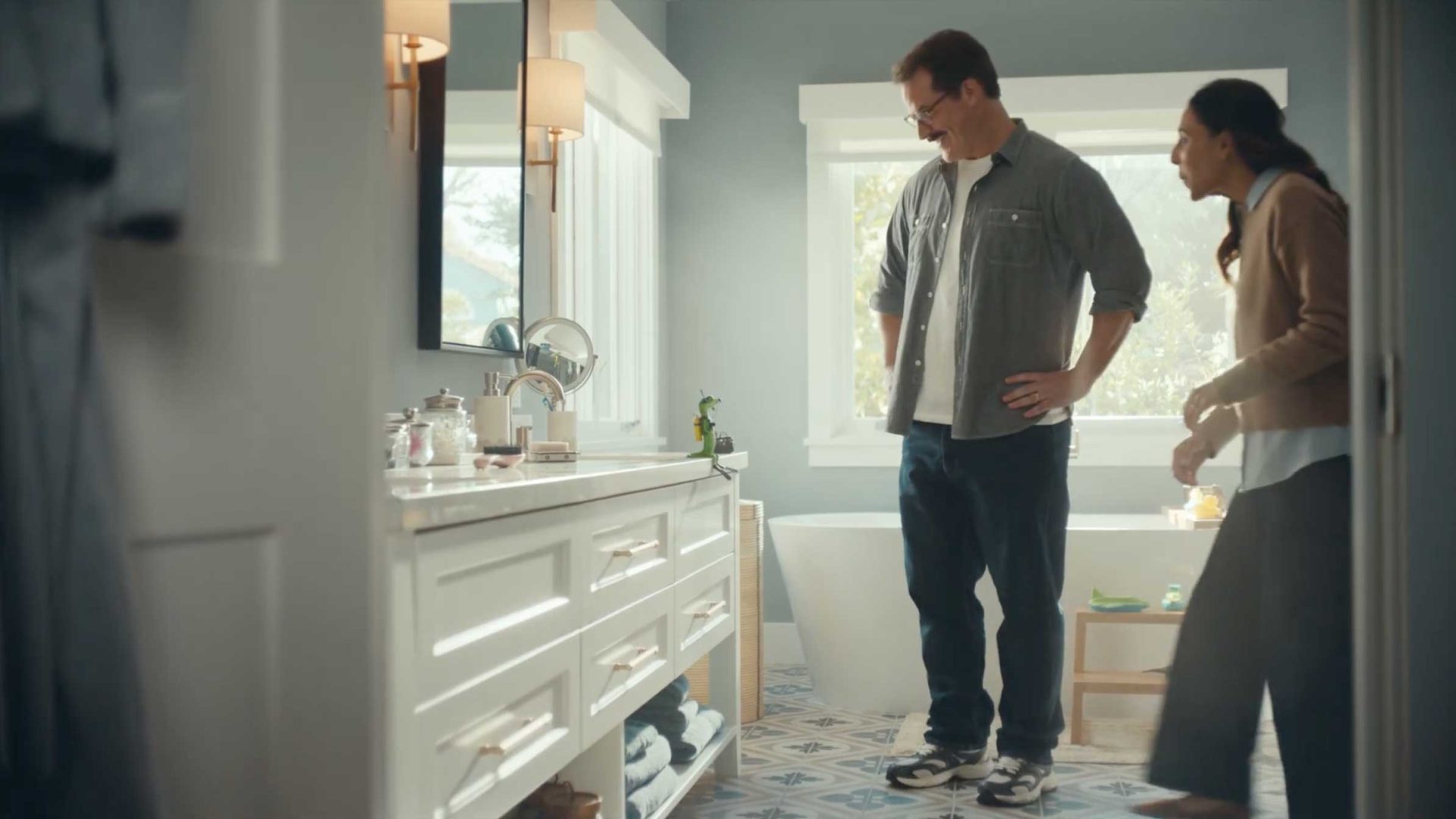 A man and woman stand and smile in a bright bathroom with a white vanity, large mirror, and bathtub. Sunlight streams in through the window, creating a cozy and inviting atmosphere reminiscent of a Framestore production—inviting viewers to imagine themselves deep diving into comfort. Frame from Stash Magazine article.