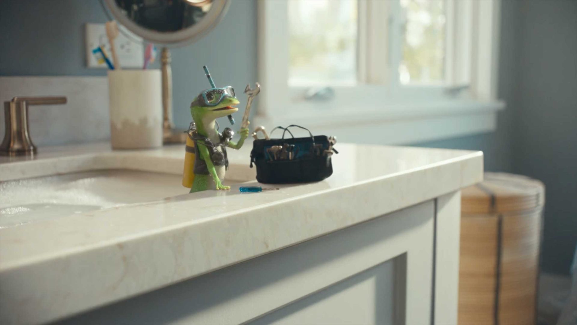 A toy lizard wearing snorkeling gear stands on a bathroom sink holding a wrench, next to a small black tool bag. The sink is overflowing with water in this playful scene inspired by the GEICO Gecko Dive commercial with VFX by Framestore. Frame from Stash Magazine article.