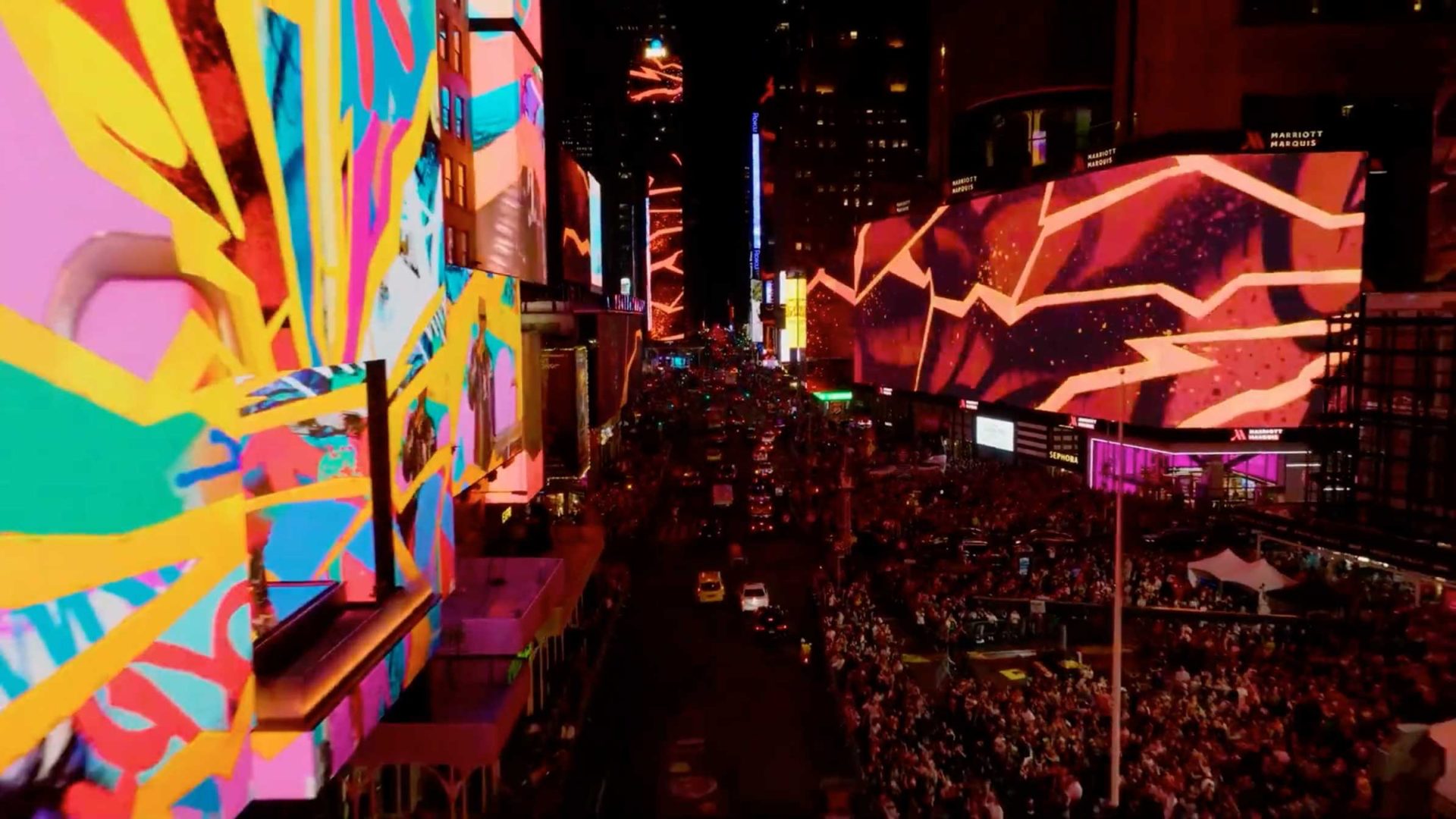 A vibrant, colorful view of Times Square at night with large digital billboards displaying abstract art by Golden Wolf, bright lights illuminating the street, and crowds of people gathered along the sidewalks. Frame from Stash Magazine article.